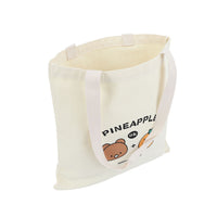 Off-white canvas tote bag with pineapple-bear logo, wide straps, side view