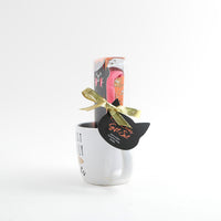 White ceramic mug with cat-shaped gift tag, paper box shows pink socks, gold ribbon