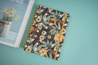 Hardcover spiral notebook with dark floral cover and gold ring binding, angled view.