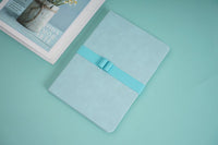 A5 notebook with light green cover and teal elastic band, closed and angled.