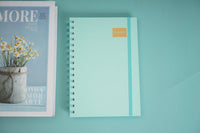 A5 daily planner notebook with turquoise cover, spiral coil binding, and gold label