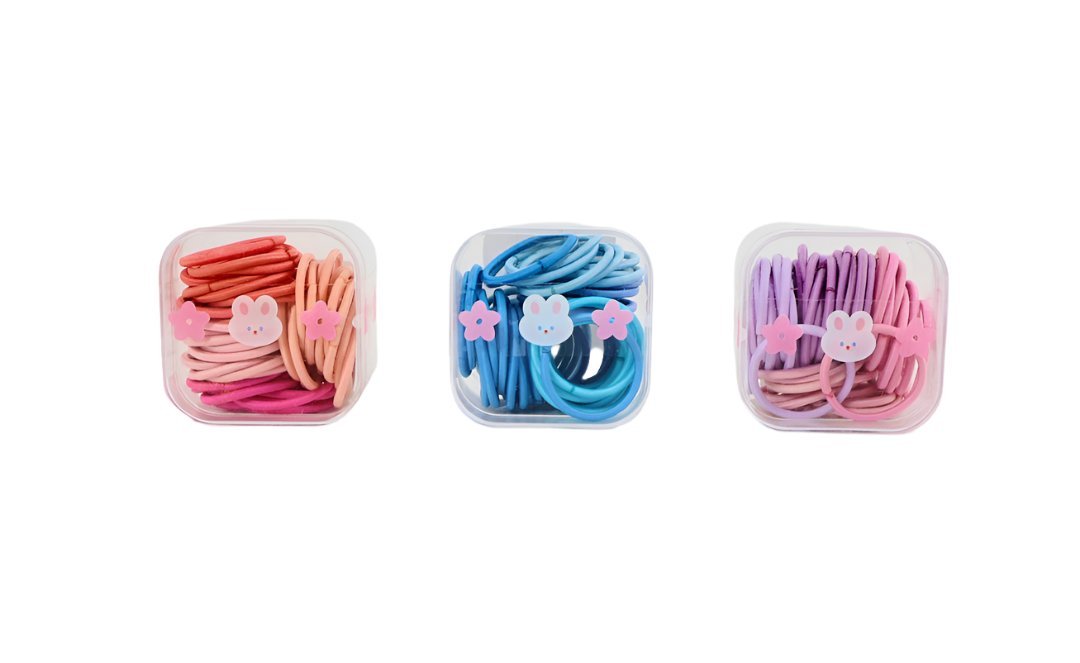 Three soft hair tie sets in clear plastic boxes: pink, blue, and purple with charms