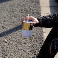 Ceramic coffee cup with lid and white straw, gold lettered top, held in hand