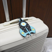 Blue rocket-shaped kids luggage tag with black strap on a white suitcase