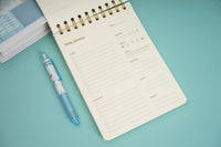 Open wood-grain coil notebook with planner layout and gold spiral rings, plus pen