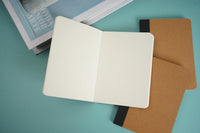 Kraft paper notebook opened to white pages, showing interior leaves and brown back cover.